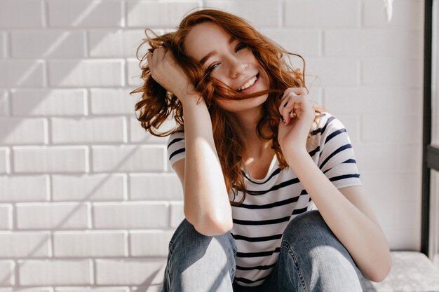 Relaxed well-dressed girl sitting. Stunning caucasian woman playing with her ginger hair.