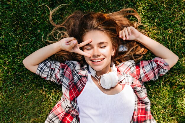 Relaxed white girl lying on lawn with peace sign. Overhead shot of pretty good-humoured woman.