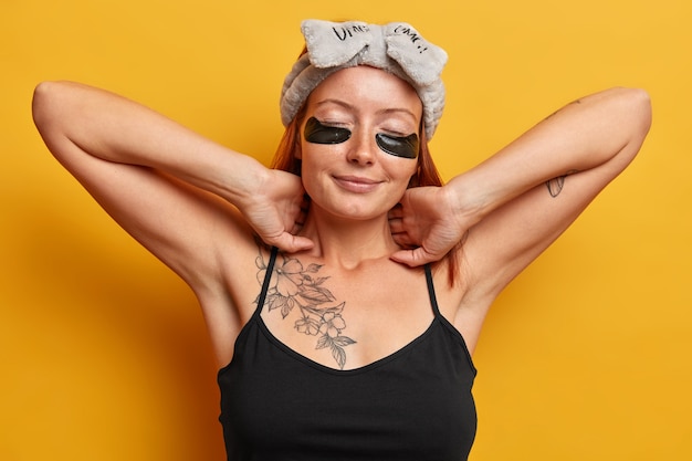 Relaxed woman stretches, puts on collagen moisturizing patches under eyes, touches tenderly neck wears hairband and black t shirt isolated over yellow wall. Facial skin care and beauty treatment