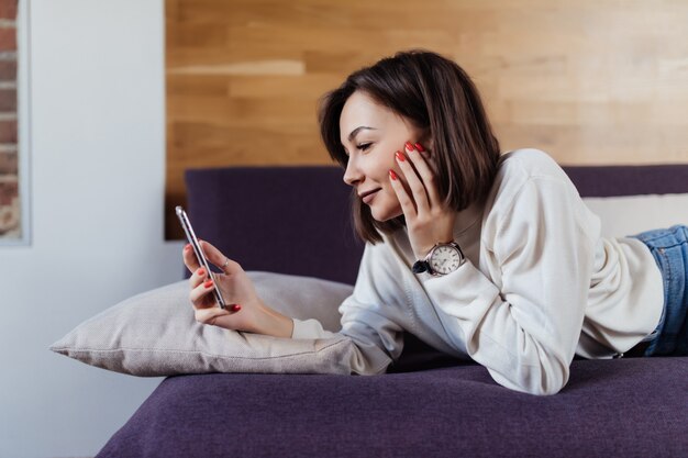自宅のベッドに横になっているスマートフォンを使用してリラックスした女性