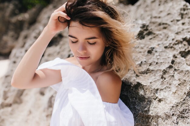 Relaxed woman in vintage white dress standing on mountain. Ecstatic dark-haired woman with tanned skin posing near rock.