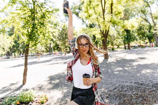 Relaxed woman with cup of coffee funny dancing in park. Debonair blonde lady in glasses enjoying music on nature in morning.