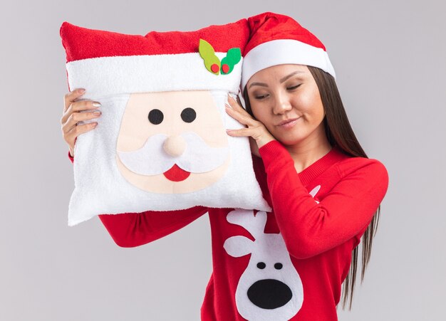 Relaxed young asian girl wearing christmas hat with sweater holding christmas pillow isolated on white background