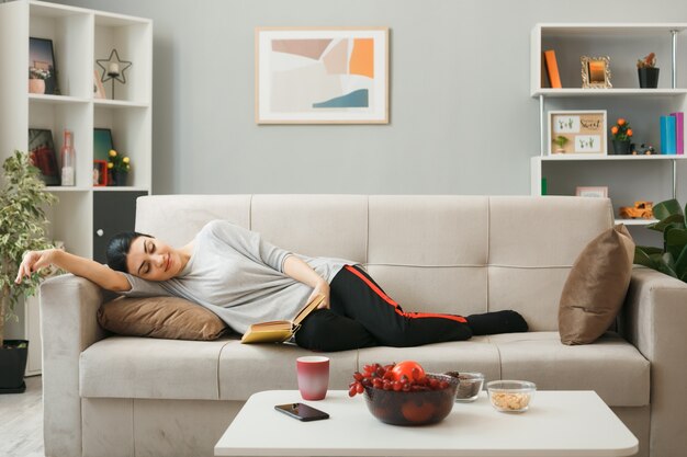 Relaxed young girl reading book lying on sofa behind coffee table in living room