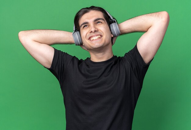 Relaxed young handsome guy wearing black t-shirt and headphones holding hands on neck isolated on green wall