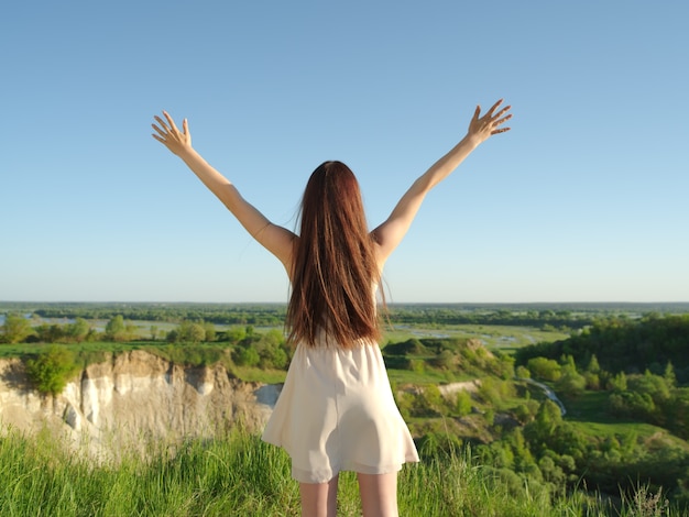 Relaxed young happy woman with raised arms outdoors in nature. Young girl stands with her arms raised up to the sky.  Peaceful girl standing by a cliff enjoying summer. - outdoors