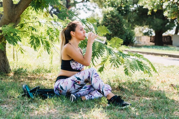 Расслабленная молодая женщина пьет воду из бутылки в саду