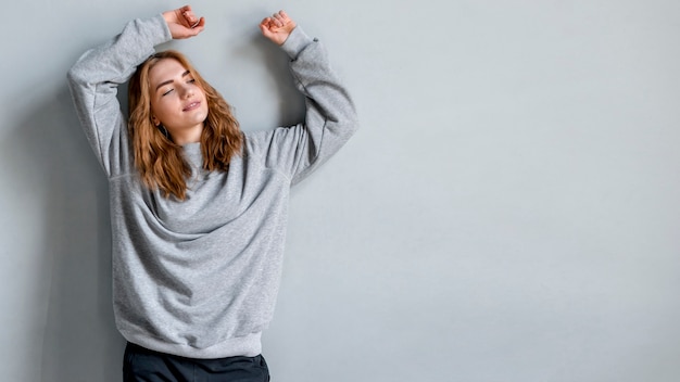 Relaxed young woman leaning on grey wall with her raised hands