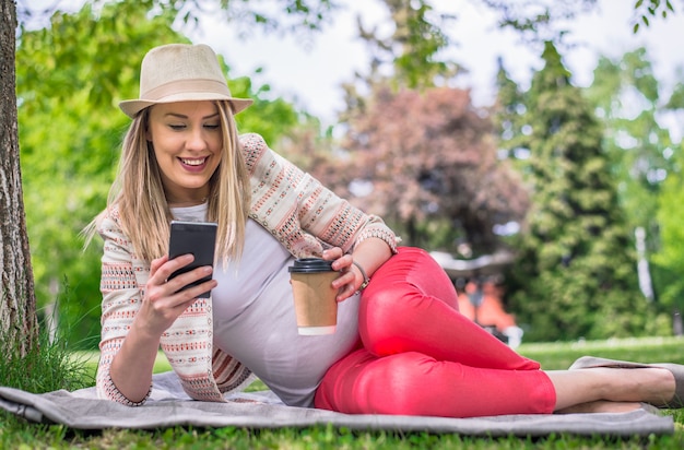 公園や庭で草の敷物に横たわって、彼女の携帯電話で彼女のテキストメッセージを読んで笑っている、ゆったりとした若い女性。草の中に横たわっている幸せな女性の全身の肖像