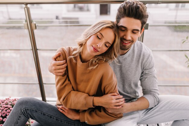 Relaxed young woman posing with husband at terrace. Handsome dark-haired man embracing girlfriend at balcony.