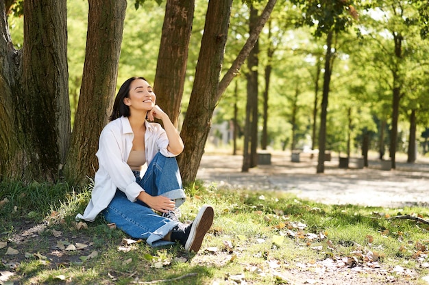 Relaxed young woman resting near tree sitting in park on lawn under shade smiling and looking happy