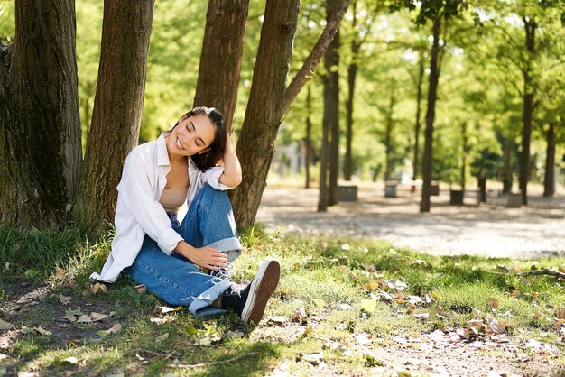 Relaxed young woman resting near tree sitting in park on lawn under shade smiling and looking happy 