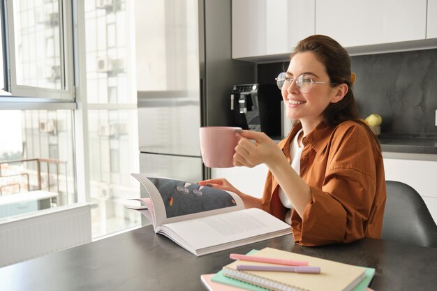 お茶やコーヒーを飲みながら本を読んでいる ⁇ 眼鏡をかぶった幸せな若い女性が家でリラックスしている日