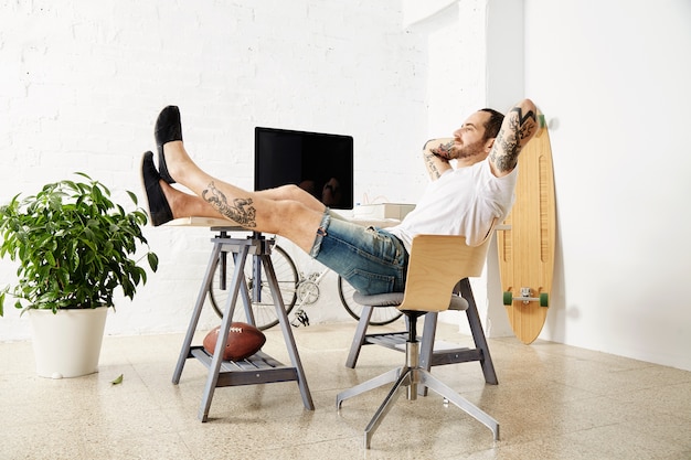 Free photo relaxing tattoed freelancer dreamily looking in window while taking break from work, with his legs on table inside big white loft