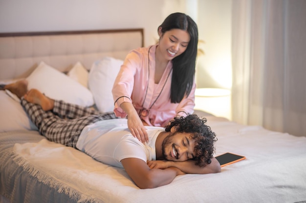 Relaxing. Young adult long haired asian woman in pink pajamas sitting giving back massage to lying smiling indian man on bed in bedroom