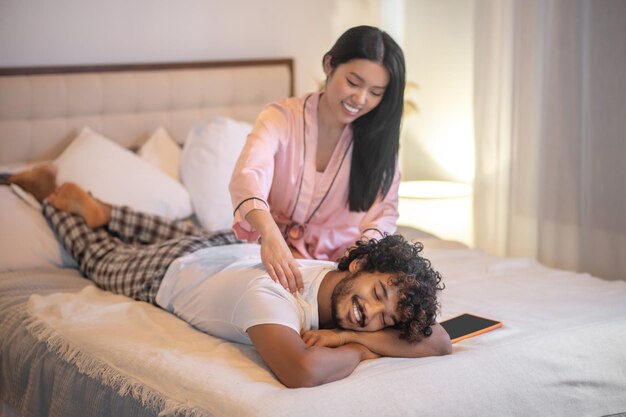 Расслабляющий. Молодая взрослая длинноволосая азиатка в розовой пижаме сидит и делает массаж спины лежащему улыбающемуся индийскому мужчине на кровати в спальне
