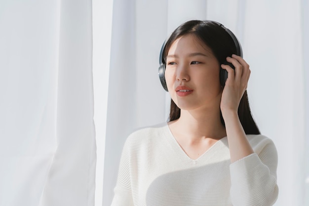 Relex and peaceful asian woman enjoy weekend with music headphone morning sunlight near window home background