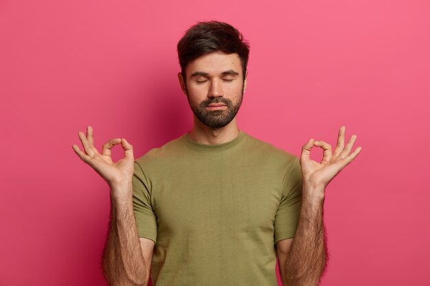 Relieved bearded young man relaxes during meditation, keeps eyes closed, spreads palms sideways in nirvana, wears casual t shirt, practices yoga, inhales fresh air, isolated on pink  wall