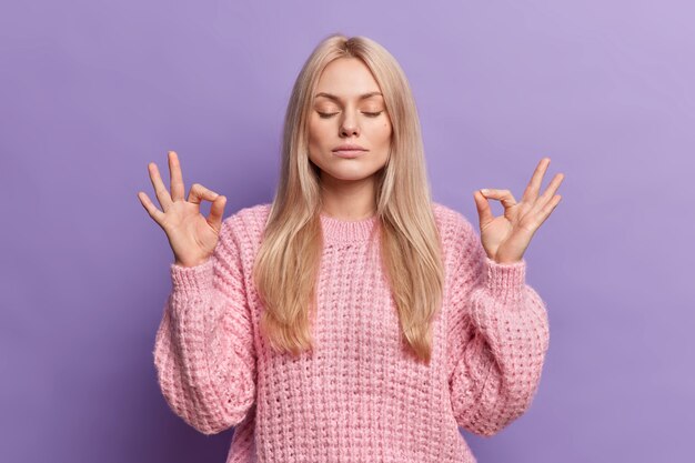 Relieved calm blonde woman searches peace inside makes mudra gesture reaches nirvana and breathes deeply with closed eyes 