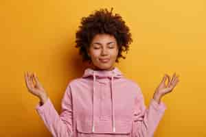 Free photo relieved ethnic woman stands in lotus pose, tries to meditate during quarantine or lockdown, reaches nirvana, does yoga, keeps eyes closed, dressed in sweatshirt. mental health, relaxation, lifestyle