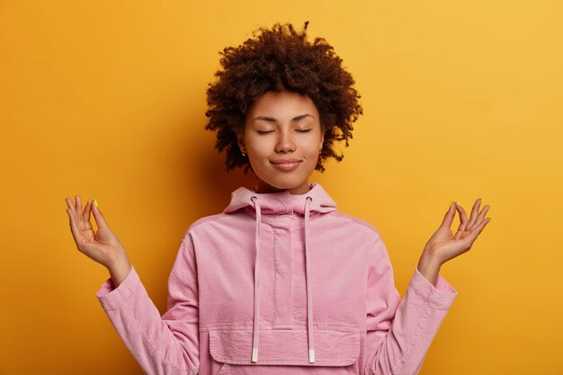 Relieved ethnic woman stands in lotus pose, tries to meditate during quarantine or lockdown, reaches nirvana, does yoga, keeps eyes closed, dressed in sweatshirt. Mental health, relaxation, lifestyle