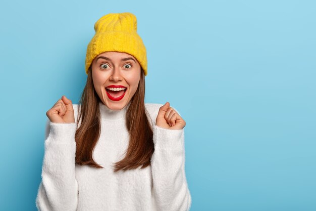 Relieved triumphing woman tilts hands in cheer, clenches fists, wears red lipstick, has dark straight hair, wears fashionable clothing, isolated over blue background