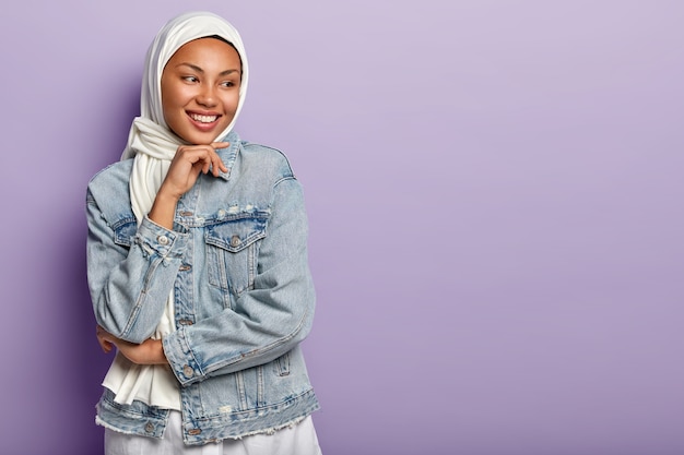 Religious Arabic woman has cheerful expression, covers head with white hijab, wears denim jacket, holds chin, looks away, stands against purple wall. People, ethnicity and faith concept