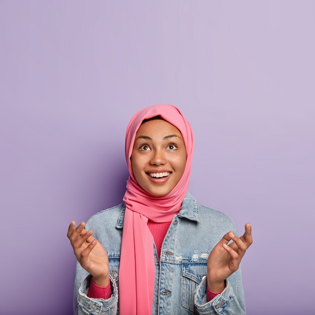 Free photo religious cheerful woman has islamic views, raises palms, prays and looks hopefully upwards