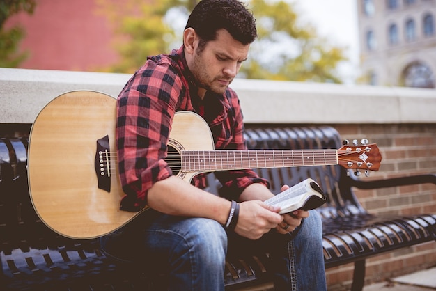 A religious man with a guitar reading the bible outdoors