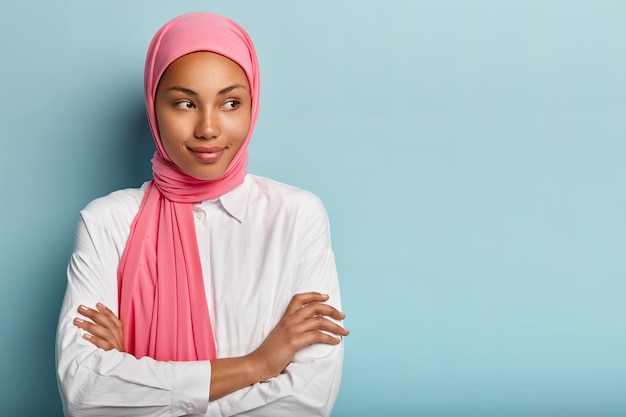 宗教的に満足している女性モデルは、腕を組んで、脇を向いて、表情を喜ばせ、頭にピンクのスカーフを着て、白いシャツを着て、深く考え、青い壁の上に立って、テキスト用のスペースをコピーします