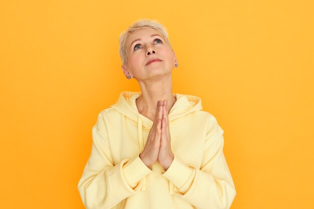 Religious unhappy woman pensioner with hopeful eyes posing isolated holding hands pressed together looking up while praying, pleading, asking God for help and guidance, being depressed in hard times