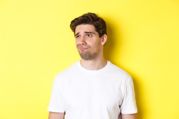 Reluctant guy in white t-shirt looking left, grimacing skeptical and displeased, standing over yellow background