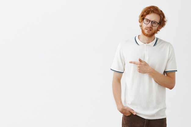 Reluctant and sad bearded redhead guy posing against the white wall with glasses