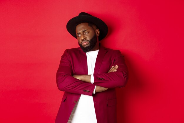 Reluctant and upset african american man looking away, cross arms on chest offended, standing distressed against red background