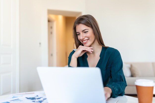 リモートジョブ技術と人々の概念は、ラップトップコンピューターとホームオフィスで働く書類を持つ若いビジネスウーマンの幸せな笑顔