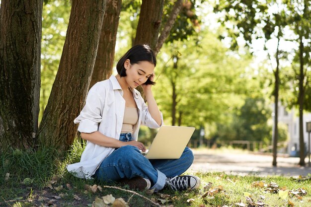 Удаленная работа, улыбающаяся азиатская студентка, делающая домашнее задание удаленно из парка, сидящая с ноутбуком возле тр