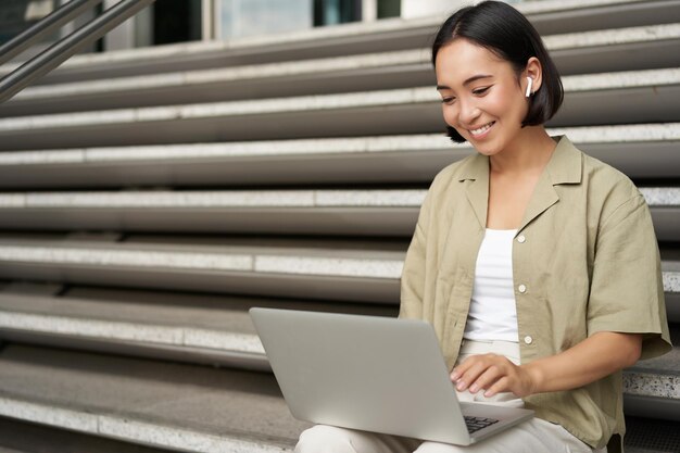 Remote worker smiling asian girl sits outdoors on street with laptop happy young woman working on co