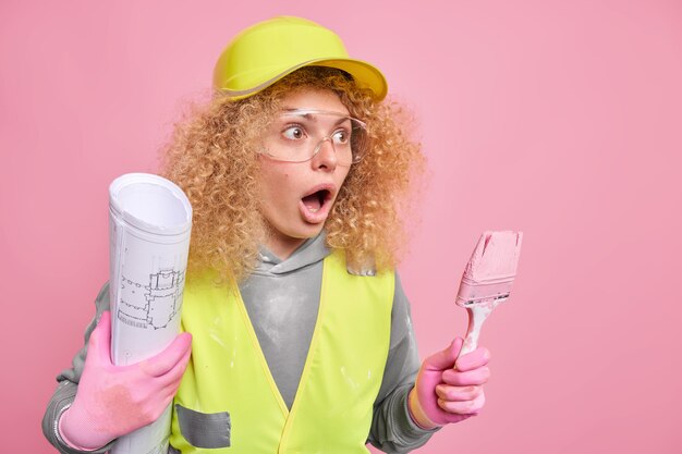 Renovation and construction concept. Scared curly haired female builder holds blueprint and painting brush looks away with stupefied expression dressed in uniform 