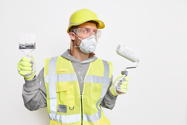 Renovation maintenance and engineering concept. Busy male labourer focused into distance with worried expression wears protective helmet safety gloves holds roller and brush follows repairing steps