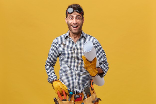 Renovation and occupation concept. Young handyman wearing protective goggles, shirt and kit full of tools holding blueprint looking with excited expression going to have rest after work