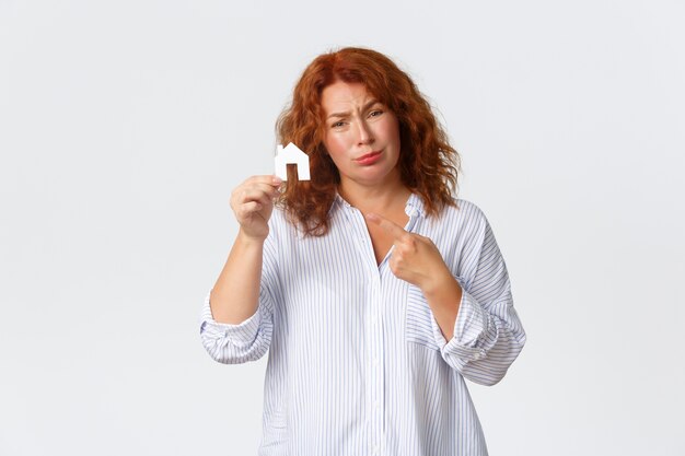Rent, buying property and real estate concept. Sad middle-aged redhead lady dreaming of having house, pointing at home cardboard and complaining not having money, need loan for buying.