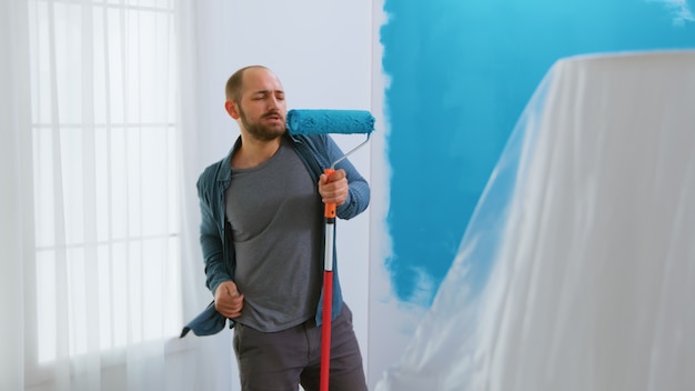 Repair man singing on roller brush with blue paint during home renovation. Dancing, construction, repairing, working. Redecoration and home construction while renovating and improving. Repair and deco