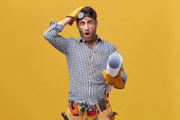 repairman in checkered shirt, gloves, protective eyeglasses, holding rolled paper having dirty face from work looking with big eyes and opened mouth after done something wrong