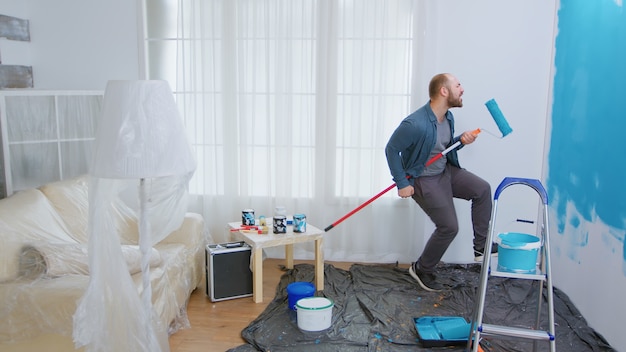 Repairman during home construction using roller brush as guitar. Guy singing while renovating house. Apartment redecoration and home construction while renovating and improving. Repair and decorating.