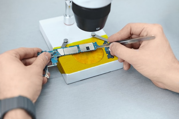 顕微鏡下で携帯電話を修理しながら、ピンセットを使用してプリント回路基板の電子部品を保持する修理工