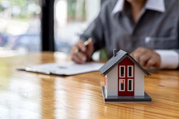 小さな​オレンジ色​の​家​の​レプリカ​が​テーブル​の​上に​あり​、​住宅​プロジェクト​の​営業​担当者​は​、​彼​が​維持している​プロジェクト​で​家​を​予約する​顧客​の​売買​契約​を​起草しています​。​不動産​取引​の​概念
