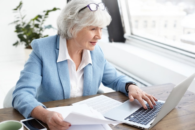 Reputable 65 year old female executive in stylish bkue suit enjoying wireless high speed internet connection while using laptop, analyzing accounts, holding papers in her hand, looking at screen