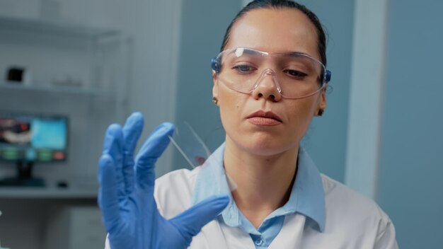 Research scientist with gloves holding blood sample on glass in laboratory. Biology doctor analyzing plasma dna for medical discovery in pharmaceutical industry. Science equipment on desk