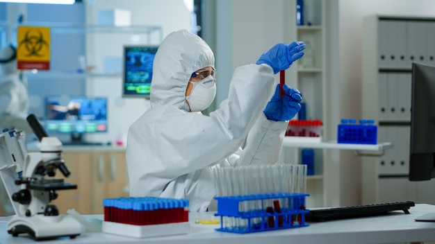 Researcher in coverall holding test tubes with blood sample for new treatment in medical laboratory. Team of doctors examining virus evolution using high tech for vaccine development against covid19