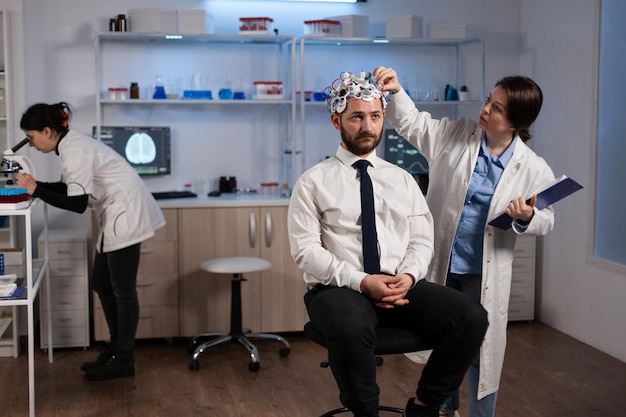 現代の実験室での医学実験中に男性患者の脳活動を監視する脳波ヘッドセットを調整する研究者の神経内科医の女性。神経系を分析する科学者の医師。医療サービス